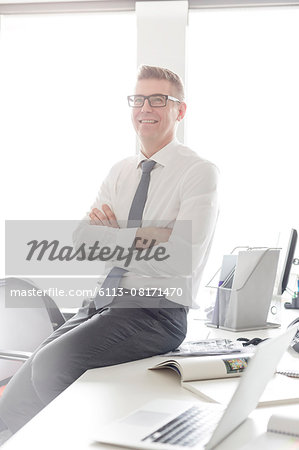 Portrait confident businessman with eyeglasses sitting on desk with arms crossed