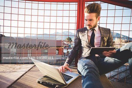 Stylish young man sitting on footbridge using laptop and digital tablet