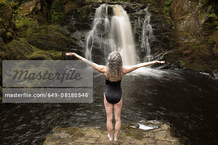 Rear view of mature woman practicing with open arms in front of waterfall