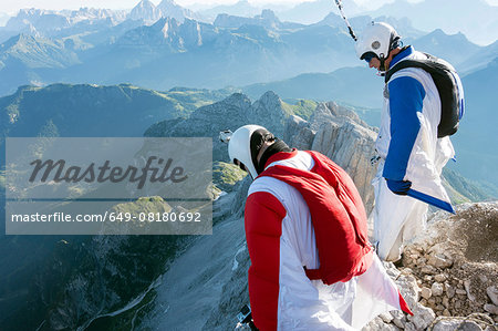 Two male BASE jumpers standing on edge of mountain looking down, Dolomites, Italy