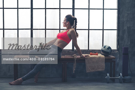 Sitting on a bench by the window in a loft gym, a fit woman in workout gear is listening to music while looking out the window.