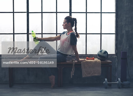 An athletic woman wearing workout gear is sitting relaxing on a wooden bench in a loft gym. Having just finished her yoga workout, she is relaxing and holding a bottle of water.