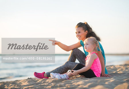 A smiling young mother points into the distance while talking to her young daughter. They have both just gone for a little run along the water's edge and are now enjoying the sunset together.