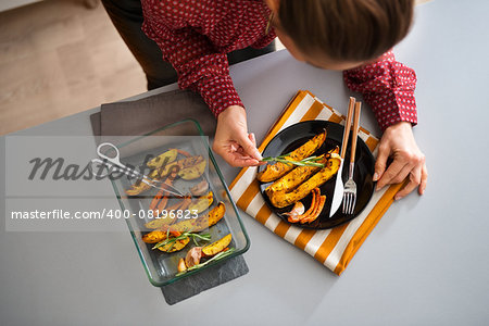 A dish of roasted autumn vegetables has just come out of the oven. An elegant woman adds the final touch of fresh rosemary to the plate as decoration.