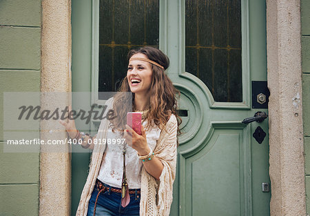 Happy trendy hipster woman with mobile phone in jeans shorts, knitted shawl and white blouse standing outdoors against olive wooden door in old town