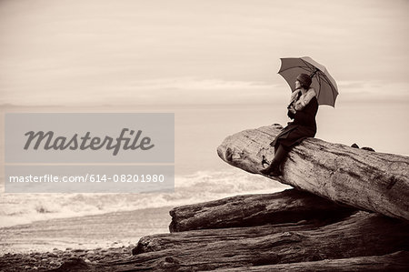 Woman with umbrella sitting on large driftwood tree trunk on beach