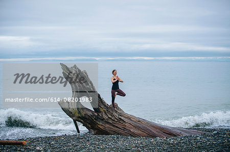 Mature woman practicing yoga tree pose on large driftwood tree trunk at beach