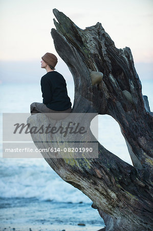 Woman sitting looking out from large driftwood tree trunk on beach