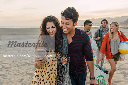 Group of friends walking along beach, young couple looking at smartphone