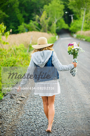 Rear view of bare footed young woman walking on rural gravel road carrying bunch of flowers