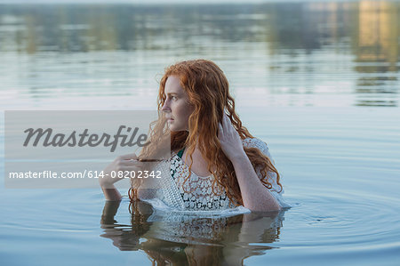 Head and shoulders of young woman with long red hair in lake looking sideways