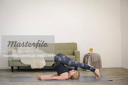 A blonde woman in a black leotard and leggings on a yoga mat with her legs bent down over her head.