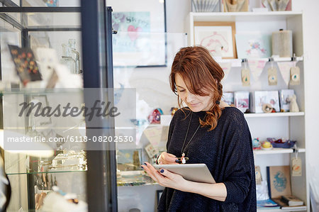 A mature woman using a digital tablet, using the touch screen, stock-taking in a small gift shop. A mature woman using a digital tablet, using the touch screen, stock-taking in a small gift shop.