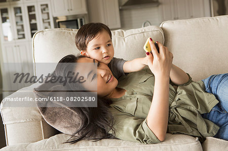Woman lying on a sofa, smiling, cuddling with her young son and looking at a cell phone.