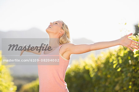 Smiling blonde woman standing in front of grapevine with eyes closed and arms outstretched