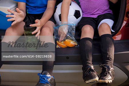 Waist down of boy and younger sister sitting in car boot eating oranges on football practice break