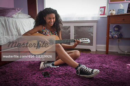 Girl sitting on bedroom floor playing guitar looking away smiling