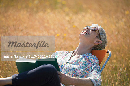 Senior woman reading book and laughing with head back in sunny field Senior woman reading book and laughing with head back in sunny field