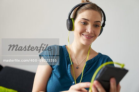 Beautiful woman relaxes on sofa and watches podcast on mobile phone, listening with green big earphones. Copy space