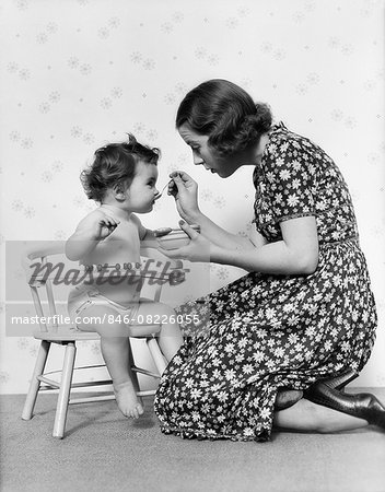 1930s KNEELING MOTHER FEEDING BABY SITTING IN CHAIR