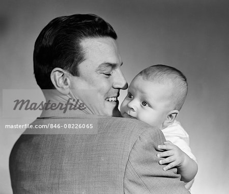 1950s PROUD SMILING MAN FATHER BACK TOWARD CAMERA HOLDING BABY SON FACE TO CAMERA