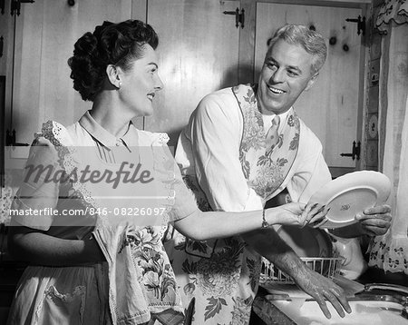 1950s SMILING HUSBAND IN APRON IN KITCHEN WASHING DISHES AS HAPPY WIFE DRIES