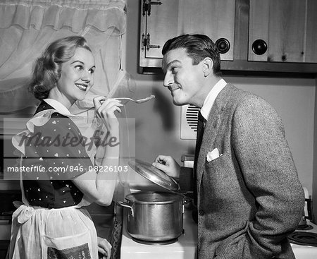 1950s SMILING HOUSEWIFE AT STOVE GIVING HAPPY HUSBAND TASTE OF HER COOKING