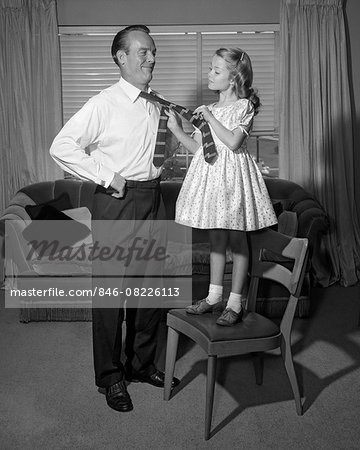 1950s CONFIDENT LITTLE GIRL STANDING ON CHAIR TYING NECKTIE FOR HER SMILING FATHER