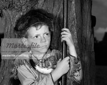 1950s BOY WEARING RACCOON SKIN HAT FRINGED WESTERN STYLE JACKET WITH POWDER HORN AND ANTIQUE RIFLE