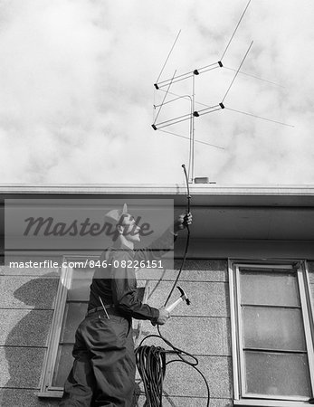 1950s 1960s MAN WORKER ON LADDER RUNNING TELEVISION CABLE TO ANTENNA ON ROOF OF HOUSE