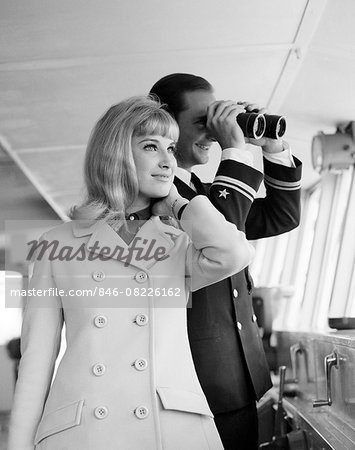 1960s MAN OFFICER IN UNIFORM USING BINOCULARS AND STYLISH BLOND WOMAN STANDING TOGETHER ON BRIDGE OF A CRUISE SHIP