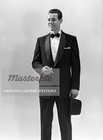 1950s PORTRAIT SMILING MAN WEARING TUXEDO HOLDING CIGARETTE AND FEDORA HAT