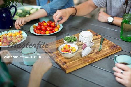 A group of people gathered around a table with plates of fresh fruits and vegetables, and a round cheese and salami on a chopping board.