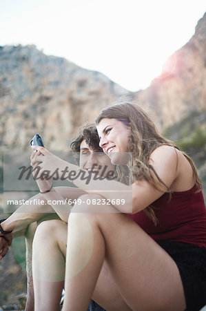 Young man and teenage sister reading smartphone texts on rocky beach, Javea, Spain