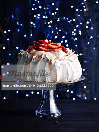 Strawberry covered pavlova on glass cake stand in front of sparkly navy blue background