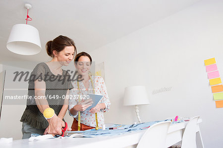 Mature women stand at desk cutting fabric looking at digital tablet