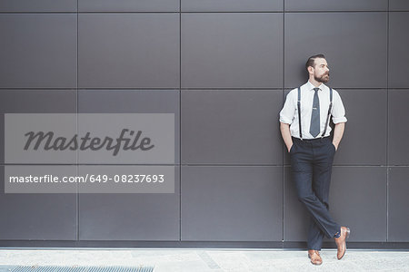 Stylish businessman with hands in pockets leaning against office wall