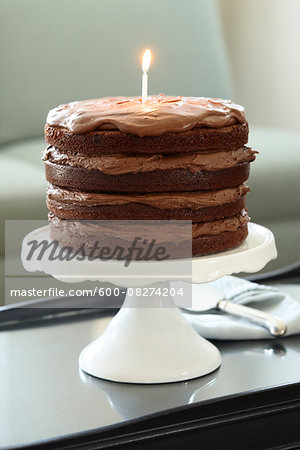 Layered Chocolate Birthday Cake with Lit Candle on Cake Stand on Coffee Table Layered Chocolate Birthday Cake with Lit Candle on Cake Stand on Coffee Table