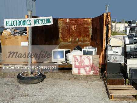 Stacks of electronics to be recycled at a landfill. One sign indicates the dropoff area, and another states that TV's are not accepted.