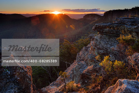 As the sunsets clipping the edge of the far plateau before sinking finally for the day, its warm golden orange rays burst forth across the valley and highlighting the lichen covered rocks and foliage at Sunset Rock, Mt Victoria