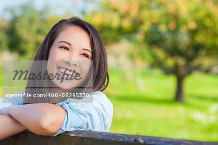 Beautiful happy Asian Eurasian young woman or girl wearing denim shirt, smiling with perfect teeth and leaning on fence in sunshine
