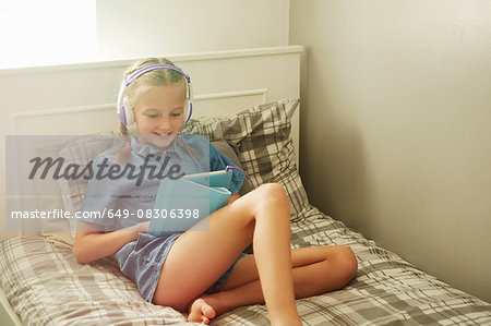 Girl sitting on bed wearing headphones looking down at digital tablet smiling