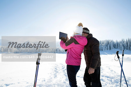 Rear view of senior couple on snowy landscape using digital tablet to take photograph of mountain range, Sattelbergalm, Tyrol, Austria