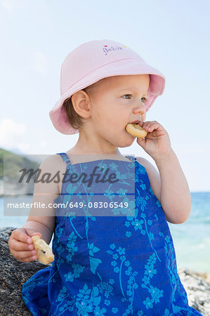 Female toddler wearing sunhat eating doughnut on beach