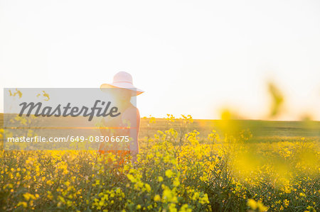 Mid adult woman in canola field wearing sunhat smiling, focus on background, lens flare