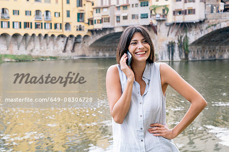 Young woman hand on hip, talking on cellular phone in front of Ponte Vecchio and river Arno looking away smiling, Florence, Tuscany, Italy