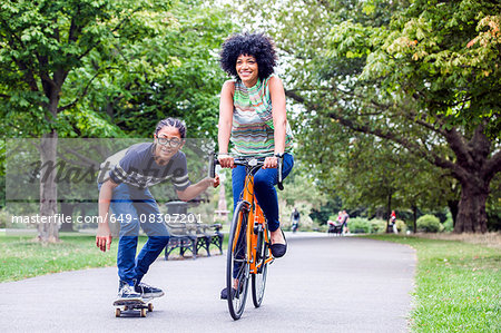 Skateboarding boy holding onto mothers bicycle in park