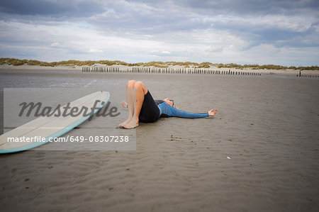 Senior woman relaxing on sand, surfboard beside her