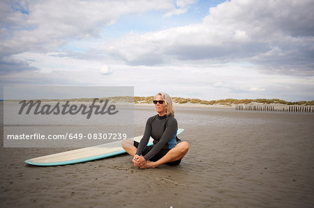Senior woman relaxing on sand, surfboard beside her
