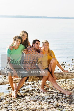 Friends sitting on driftwood next to shoreline looking at camera smiling, Schondorf, Ammersee, Bavaria, Germany